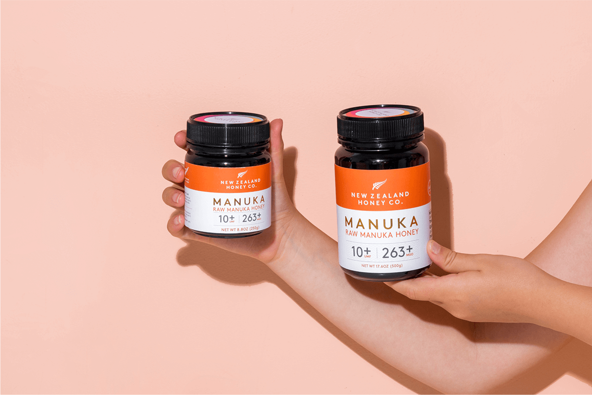 Zwei Hände halten je ein Glas Manuka Honig UMF™ 10+ MGO 263+ vor rosa Hintergrund