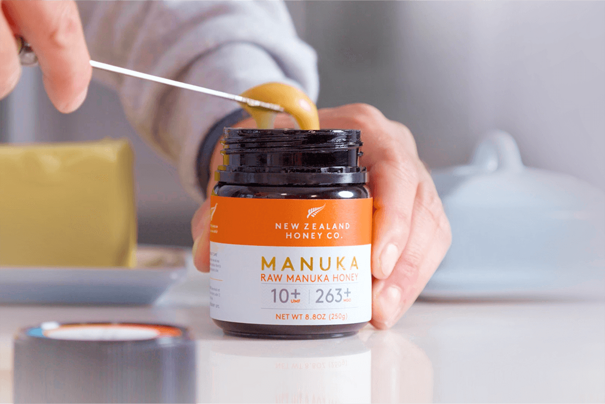 Person entnimmt Manuka Honig mit Löffel aus geöffnetem UMF™ 10+ MGO 263+ Glas – Nahaufnahme