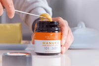 Person entnimmt Manuka Honig mit Löffel aus geöffnetem UMF™ 10+ MGO 263+ Glas – Nahaufnahme