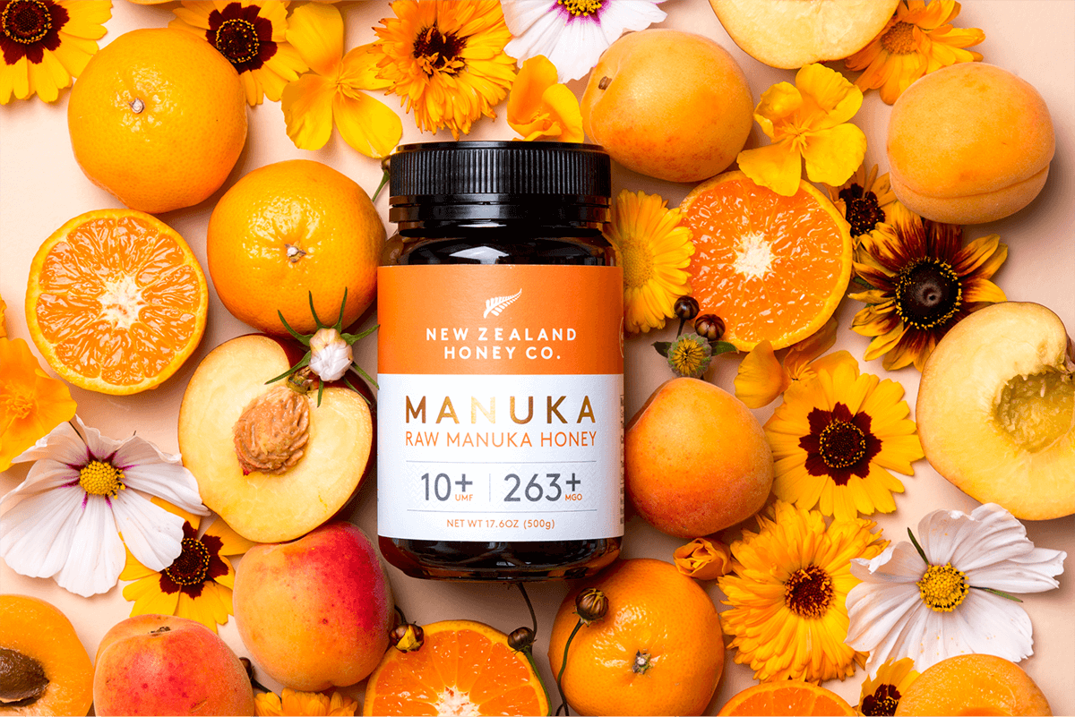 Manuka Honig UMF™ 10+ MGO 263+ Glas umgeben von Orangen, Pfirsichen und Blumen – Flat-Lay