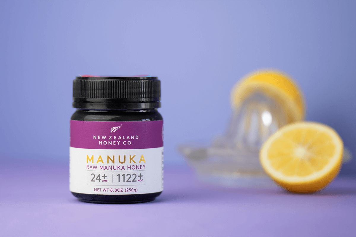Zwei Gläser Manuka Honig UMF™ 24+ MGO 1122+ mit Reagenzglas und Honigkristallen vor grünem Hintergrund