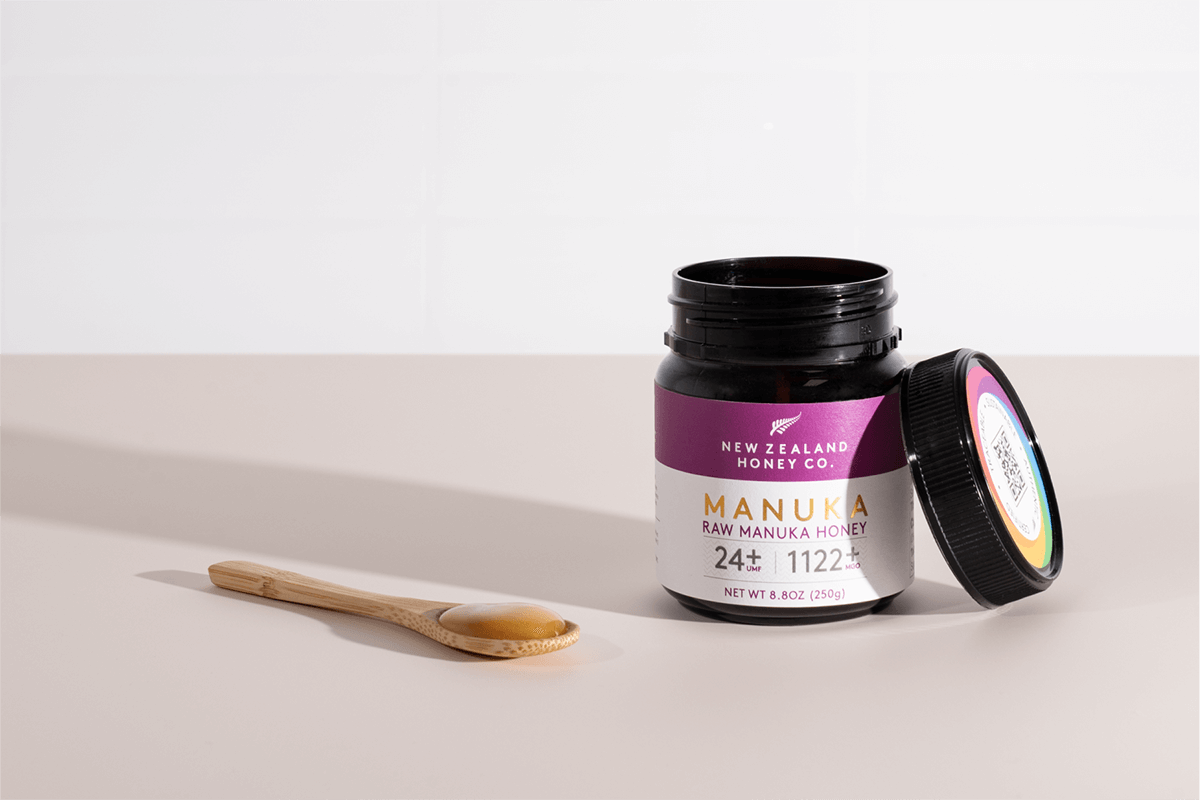 Geöffnetes Glas Manuka Honig UMF™ 24+ MGO 1122+ mit Holzlöffel auf neutralem Hintergrund