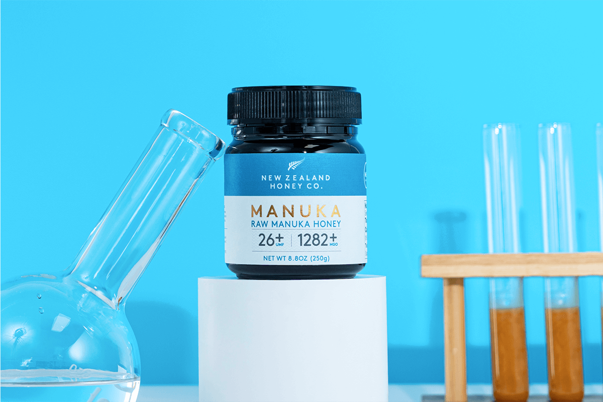 Geöffnetes Glas Manuka Honig UMF™ 26+ MGO 1282+ mit Holzlöffel auf neutralem Hintergrund