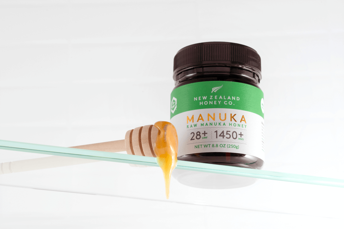 Manuka Honig UMF™ 28+ MGO 1450+ Glas mit Holz-Honiglöffel – Honig tropft auf Glasregal, weißer Hintergrund