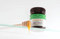 Manuka Honig UMF™ 28+ MGO 1450+ Glas mit Holz-Honiglöffel – Honig tropft auf Glasregal, weißer Hintergrund