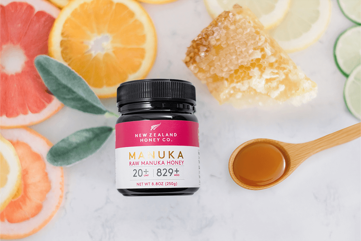 Manuka Honig UMF™ 20+ MGO 829+ Glas, Flat-Lay mit Zitrusfrüchten, Honigwabe und Holzlöffel auf Marmor