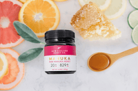 Manuka Honig UMF™ 20+ MGO 829+ Glas, Flat-Lay mit Zitrusfrüchten, Honigwabe und Holzlöffel auf Marmor