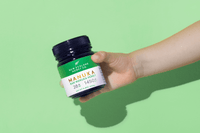 Hand hält Manuka Honig UMF™ 28+ MGO 1450+ Glas vor grünem Hintergrund