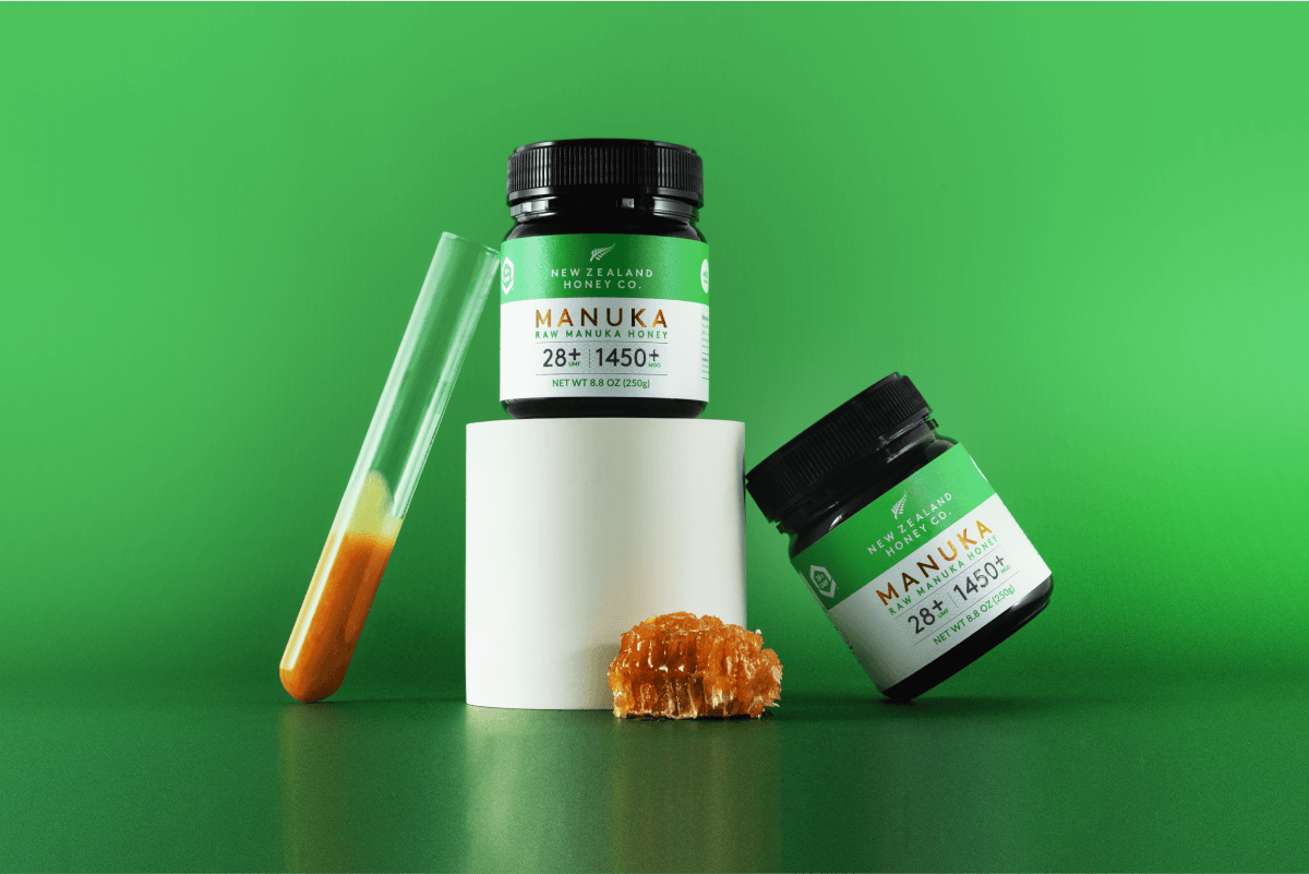 Zwei Gläser Manuka Honig UMF™ 28+ MGO 1450+ mit Reagenzglas und Honigkristallen vor grünem Hintergrund