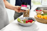 Person hält Manuka Honig UMF™ 20+ MGO 829+ über Smoothie-Bowl mit frischen Früchten in heller Küche