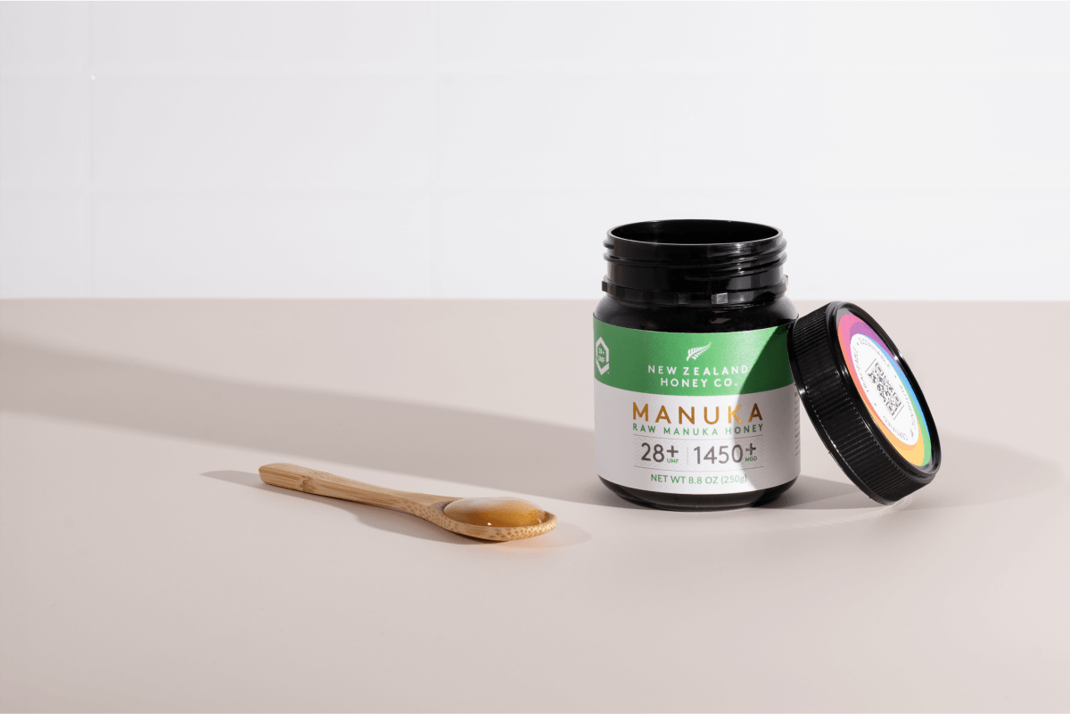 Geöffnetes Glas Manuka Honig UMF™ 28+ MGO 1450+ mit Holzlöffel auf hellem Studiohintergrund