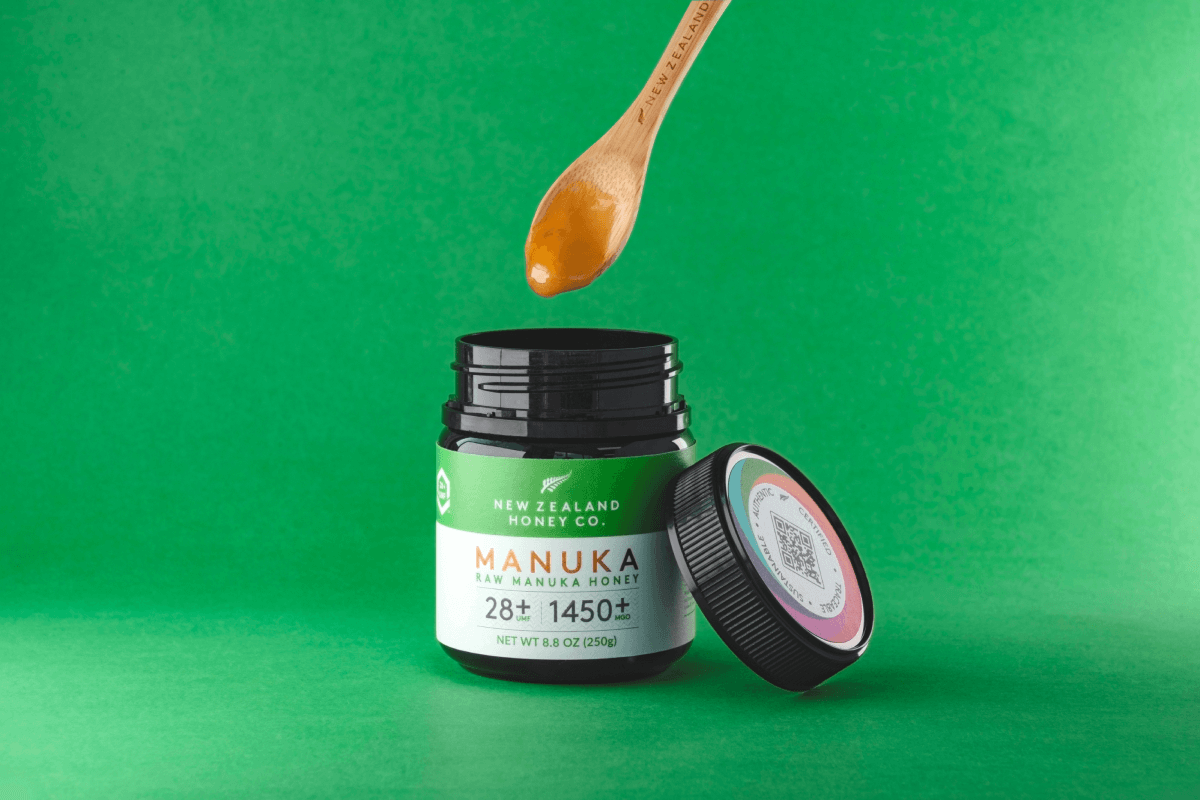 Geöffnetes Glas Manuka Honig UMF™ 28+ MGO 1450+ mit tropfendem Honiglöffel vor grünem Hintergrund